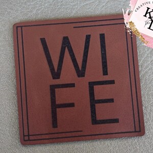 May include: A square, brown coaster with the word "WIFE" in large, black capital letters. The coaster has a thin, black border and is set against a textured, gray surface. A small logo is in the upper right corner.