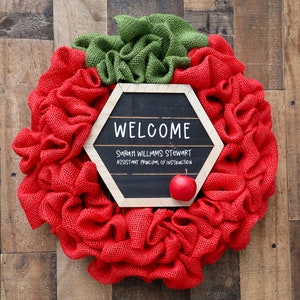 May include: A red and green burlap wreath with a hexagonal chalkboard sign that reads "Welcome, Sarah Williams Stewart, Assistant Principal of Instruction". A small red apple is attached to the sign.