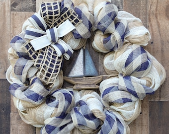 May include: A nautical themed wreath with a burlap and blue and white checkered mesh base. A wooden sailboat with a blue sail is in the center of the wreath. A blue and white ribbon with a burlap bow is on the top of the wreath.