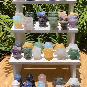 May include: A selection of small, carved gemstone frog figurines in various colours, including pink, blue, green, and clear. The figurines are displayed on a white, tiered shelf, with a wooden surface below. The frogs have a whimsical design.