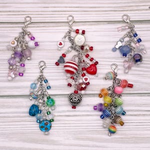 May include: Five colorful charm keychains with silver chains and various beads. The charms include a red and white striped bead, a red heart, a blue heart, a silver heart, and a rainbow of beads.