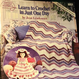 May include: A crochet pattern book titled "Learn to Crochet in Just One Day" by Jean Leinhauser. The cover features a crocheted blanket with a wavy striped pattern in shades of purple, pink, and white. A doll wearing a crocheted dress is sitting on a couch in the background.