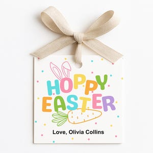 May include: A square white gift tag with the words "HOPPY EASTER" in colorful letters, a carrot illustration, and the text "Love, Olivia Collins." The tag is tied with a beige ribbon.
