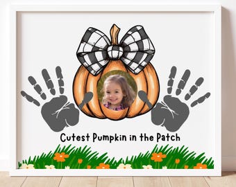 Può includere: Stampa incorniciata con un design a zucca e un fiocco a scacchi bianco e nero. La zucca ha una foto di un bambino al centro. Impronte di mani grigie affiancano la zucca, con il testo "Cutest Pumpkin in the Patch" in basso.