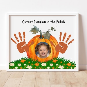 Könnte beinhalten: Gerahmter Druck mit dem Foto eines Kindes in einer orangefarbenen Kürbisillustration. Handabdrücke flankieren den Kürbis, über einem grasbewachsenen Feld mit Blumen. Der Text "Cutest Pumpkin in the Patch" ist oben auf dem Druck zu sehen.