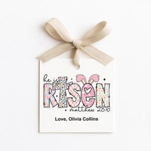 May include: A white gift tag with a beige ribbon bow. The tag features the words "he is RISEN" in colorful, patterned letters, with bunny ears above the "S". The text "matthew 28:6" and "Love, Olivia Collins" are also present.