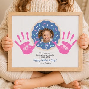 May include: A framed print featuring a child's photo inside a blue donut with sprinkles. Pink handprints flank the donut. Text reads "You donut know how much I love you! Happy Father's Day! Love, Olivia."