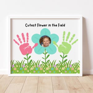 May include: A framed print featuring a child's photo in a turquoise flower design, flanked by pink and green handprints. The words "Cutest Flower in the Field" are printed above the flower. A colorful grass border is at the bottom.