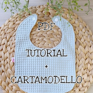 Può includere: Un bavaglino azzurro con trama a nido d'ape, fissato con due bottoni bianchi. Il bavaglino presenta il testo "PDF TUTORIAL + CARTAMODELLO" in nero. Il bavaglino è posto su un sottobicchiere rotondo intrecciato, con fogliame verde sullo sfondo.