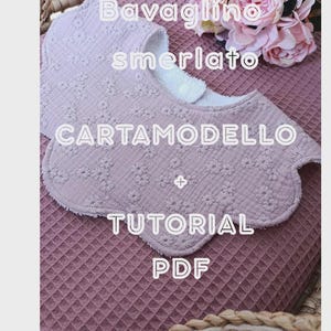 Puede incluir: Babero rosa claro con un estampado floral y bordes festoneados. El babero está hecho de una tela suave y texturizada. El texto en el babero incluye "Bavaglino smerlato CARTAMODELLO + TUTORIAL PDF".