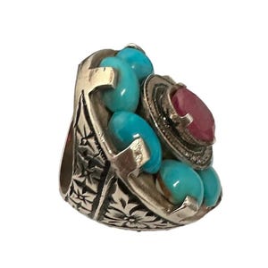 May include: A silver ring with a large red gemstone surrounded by smaller turquoise gemstones. The ring has an intricate floral design.