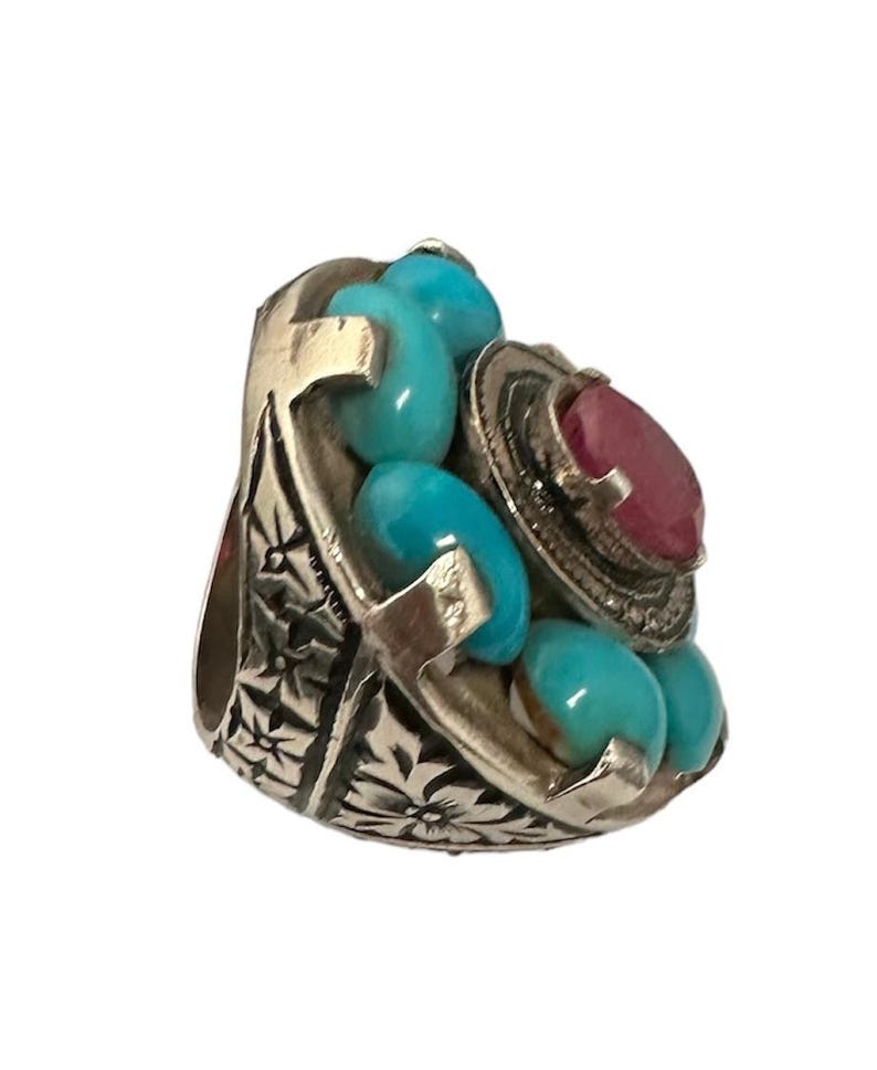 May include: A silver ring with a large red gemstone surrounded by smaller turquoise gemstones. The ring has an intricate floral design.