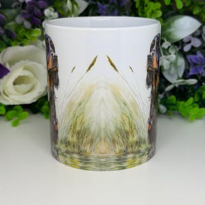 K&ouml;nnte beinhalten: Wei&szlig;e Keramiktasse mit einer detaillierten Illustration eines Hundes mit braunem und schwarzem Fell, vor dem Hintergrund von hohem Gras und Wasser. Das Design ist auf der Tasse gespiegelt, wodurch ein symmetrischer Effekt entsteht. Ein dekorativer Artikel f&uuml;r Hundeliebhaber.