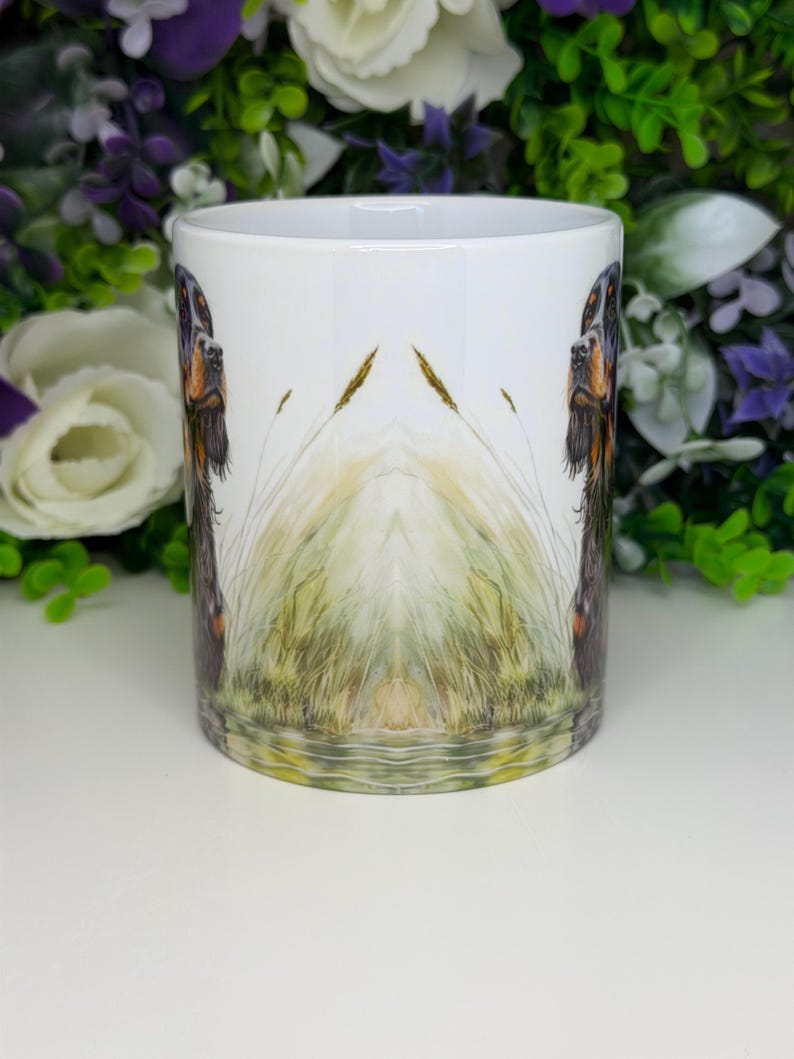 K&ouml;nnte beinhalten: Wei&szlig;e Keramiktasse mit einer detaillierten Illustration eines Hundes mit braunem und schwarzem Fell, vor dem Hintergrund von hohem Gras und Wasser. Das Design ist auf der Tasse gespiegelt, wodurch ein symmetrischer Effekt entsteht. Ein dekorativer Artikel f&uuml;r Hundeliebhaber.