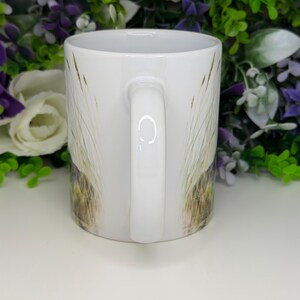 K&ouml;nnte beinhalten: Ein wei&szlig;er Keramikbecher mit einem aufgedruckten Design aus hohen, d&uuml;nnen Grashalmen in Gr&uuml;n- und Braunt&ouml;nen. Der Becher hat eine glatte, gl&auml;nzende Oberfl&auml;che, einen zylindrischen K&ouml;rper und einen geschwungenen Henkel an der Seite.