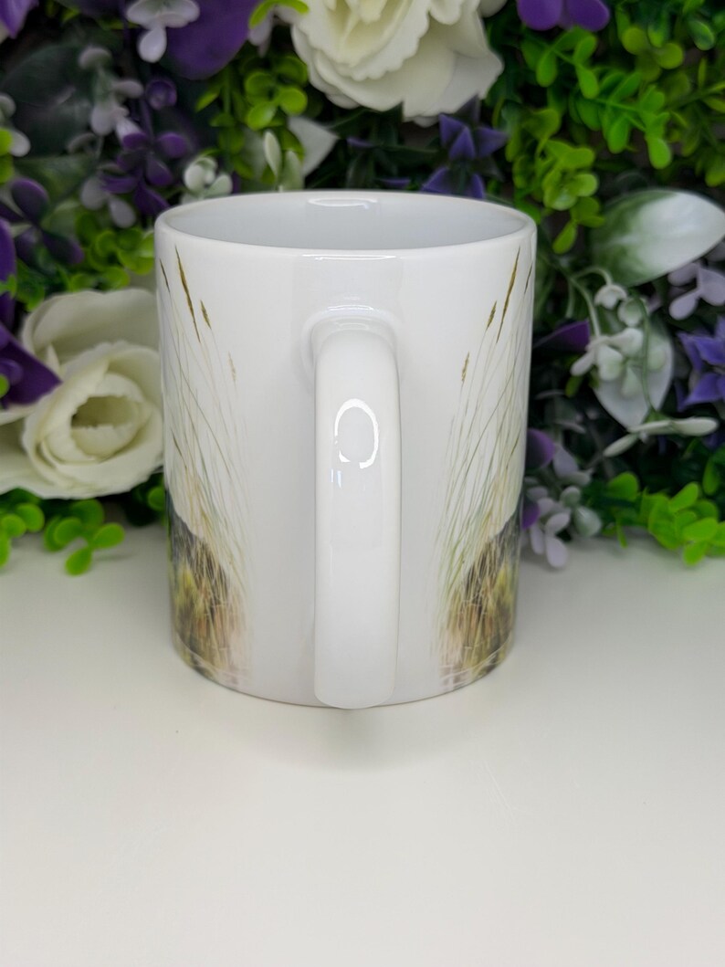 K&ouml;nnte beinhalten: Ein wei&szlig;er Keramikbecher mit einem aufgedruckten Design aus hohen, d&uuml;nnen Grashalmen in Gr&uuml;n- und Braunt&ouml;nen. Der Becher hat eine glatte, gl&auml;nzende Oberfl&auml;che, einen zylindrischen K&ouml;rper und einen geschwungenen Henkel an der Seite.