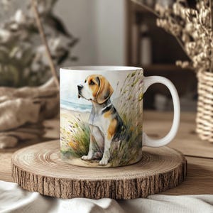 May include: White ceramic mug with a watercolour illustration of a beagle dog on a beach. The dog has brown, black, and white markings. The background shows a beach scene with grass and the sea.