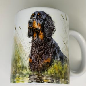 K&ouml;nnte beinhalten: Wei&szlig;e Keramiktasse mit einer detaillierten Illustration eines Gordon Setter Hundes. Der Hund hat schwarzes und braunes Fell mit einem fokussierten Blick. Der Hintergrund zeigt hohes Gras und Wasser, wodurch eine naturalistische Szene entsteht. Die Tasse hat einen wei&szlig;en Henkel.