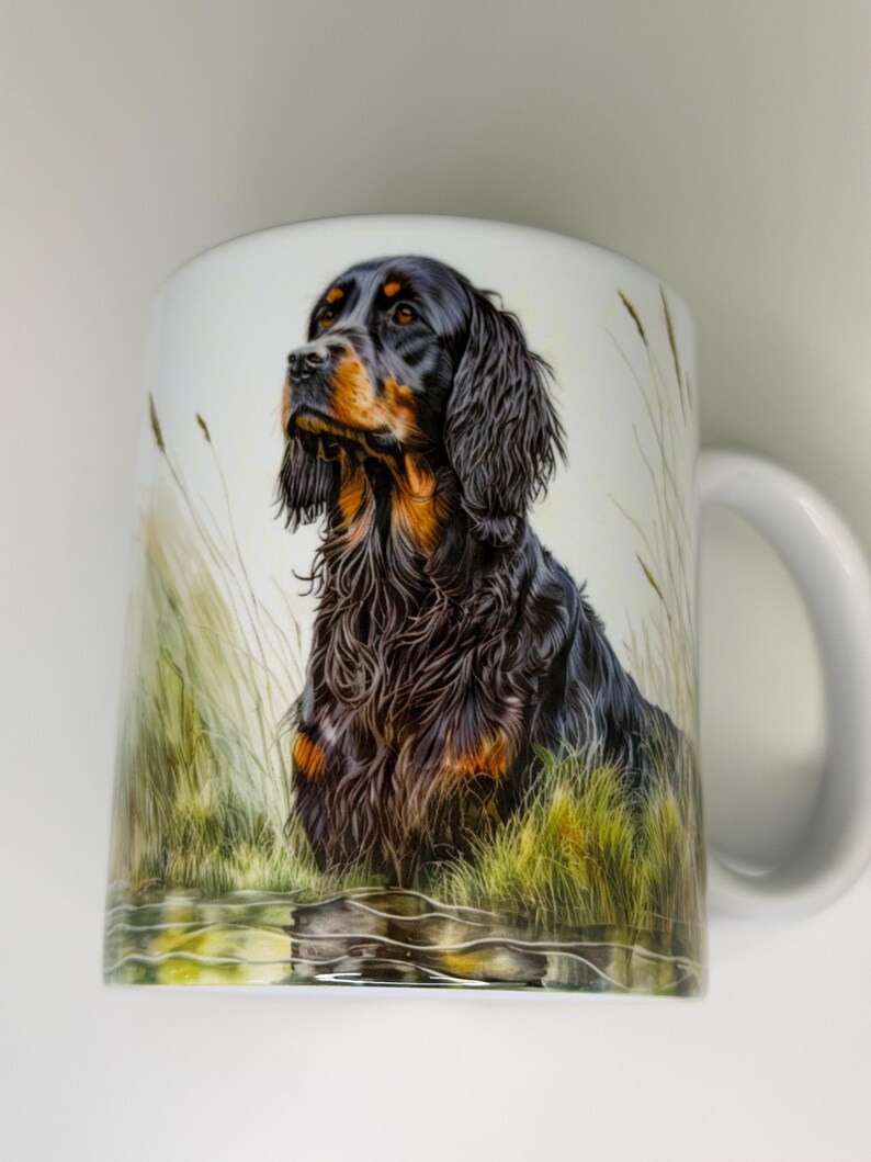 K&ouml;nnte beinhalten: Wei&szlig;e Keramiktasse mit einer detaillierten Illustration eines Gordon Setter Hundes. Der Hund hat schwarzes und braunes Fell mit einem fokussierten Blick. Der Hintergrund zeigt hohes Gras und Wasser, wodurch eine naturalistische Szene entsteht. Die Tasse hat einen wei&szlig;en Henkel.
