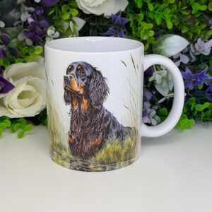 K&ouml;nnte beinhalten: Wei&szlig;e Keramiktasse mit einer detaillierten Illustration eines Gordon Setter-Hundes in einem grasbewachsenen Feld. Der Hund hat schwarzes und braunes Fell. Die Tasse hat einen wei&szlig;en Henkel und ist von gr&uuml;nem und violettem Laub umgeben.
