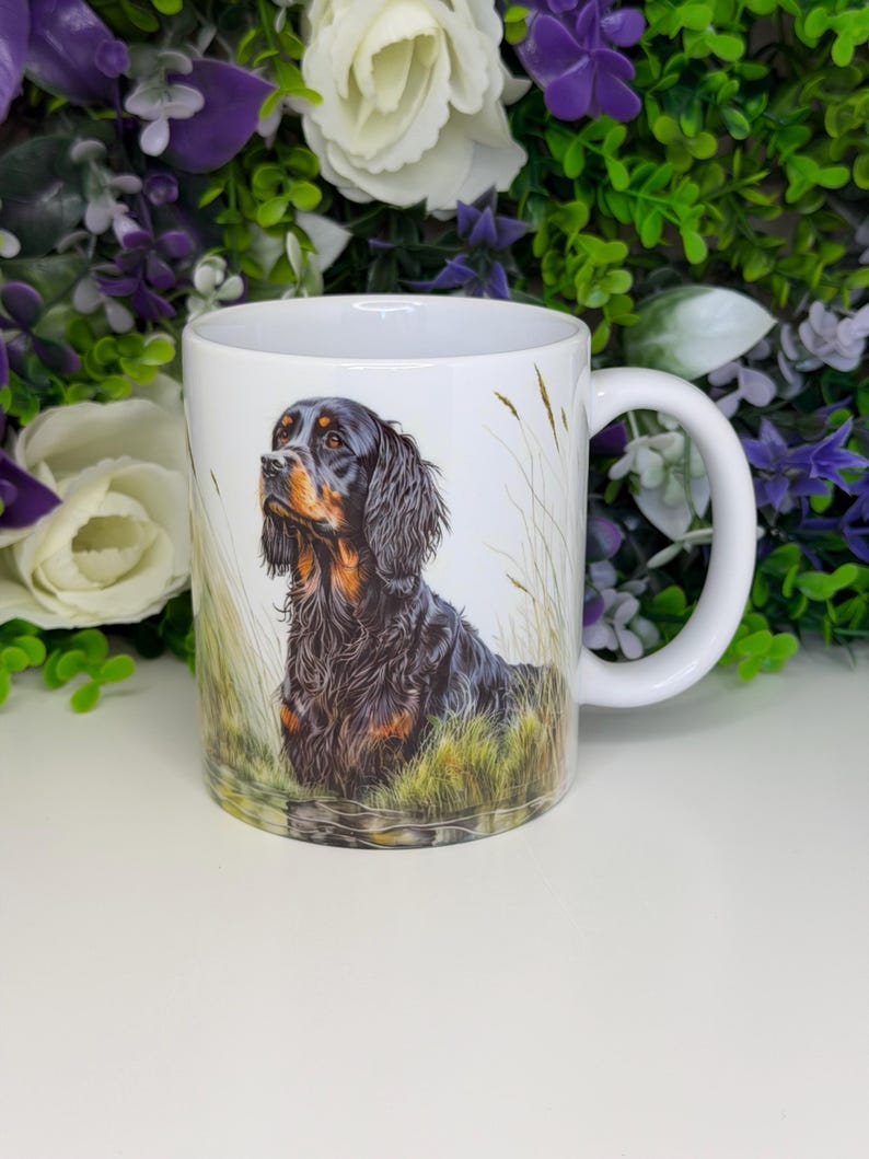 K&ouml;nnte beinhalten: Wei&szlig;e Keramiktasse mit einer detaillierten Illustration eines Gordon Setter-Hundes in einem grasbewachsenen Feld. Der Hund hat schwarzes und braunes Fell. Die Tasse hat einen wei&szlig;en Henkel und ist von gr&uuml;nem und violettem Laub umgeben.