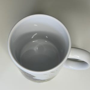 K&ouml;nnte beinhalten: Ein wei&szlig;er Keramikbecher von oben betrachtet, der einen glatten, hohlen Innenraum und einen nach rechts ragenden, geschwungenen Henkel zeigt. Die Au&szlig;enfl&auml;che weist einen teilweisen Grafikdruck mit gedeckten T&ouml;nen auf.