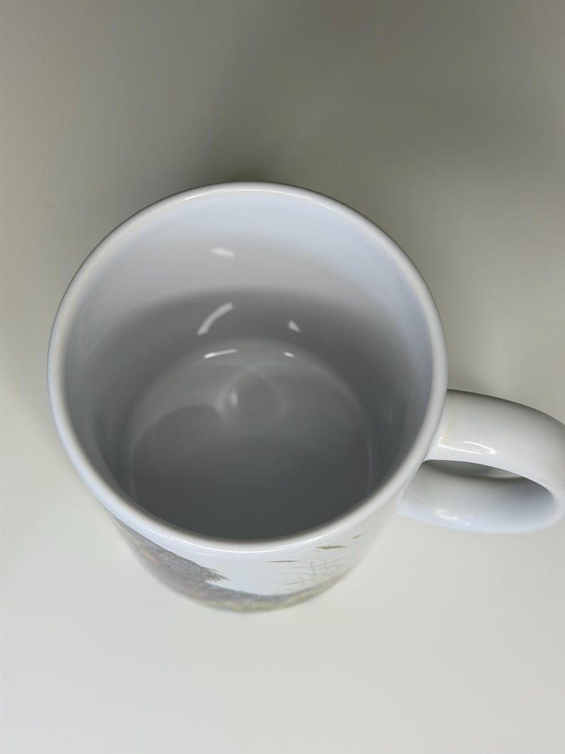 K&ouml;nnte beinhalten: Ein wei&szlig;er Keramikbecher von oben betrachtet, der einen glatten, hohlen Innenraum und einen nach rechts ragenden, geschwungenen Henkel zeigt. Die Au&szlig;enfl&auml;che weist einen teilweisen Grafikdruck mit gedeckten T&ouml;nen auf.