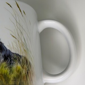 K&ouml;nnte beinhalten: Ein wei&szlig;er Keramikbecher mit einer detaillierten Illustration eines langhaarigen schwarzen Hundes, der teilweise zwischen hohen, goldgr&uuml;nen Grashalmen verborgen ist. Der Becher hat einen glatten, geschwungenen Henkel an der rechten Seite.