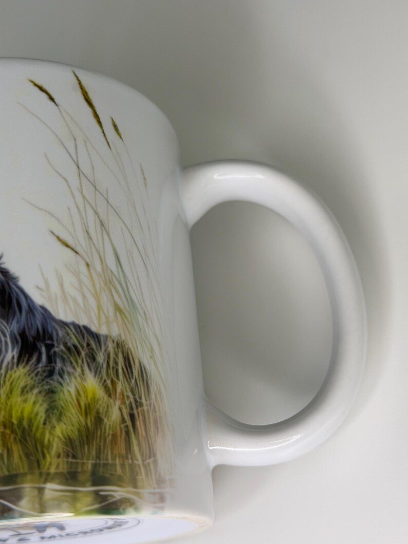 K&ouml;nnte beinhalten: Ein wei&szlig;er Keramikbecher mit einer detaillierten Illustration eines langhaarigen schwarzen Hundes, der teilweise zwischen hohen, goldgr&uuml;nen Grashalmen verborgen ist. Der Becher hat einen glatten, geschwungenen Henkel an der rechten Seite.