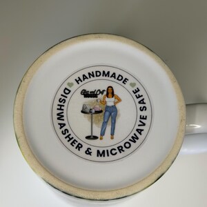 K&ouml;nnte beinhalten: Der Boden einer wei&szlig;en Keramiktasse zeigt ein kreisf&ouml;rmiges Logo mit dem Text HANDMADE und DISHWASHER & MICROWAVE SAFE. Das Logo enth&auml;lt eine Illustration einer Person, die neben einem Tisch mit einem Laptop steht, sowie den Text Glass and Craft HOUSE.