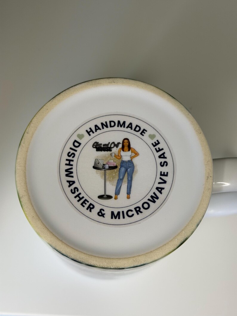K&ouml;nnte beinhalten: Der Boden einer wei&szlig;en Keramiktasse zeigt ein kreisf&ouml;rmiges Logo mit dem Text HANDMADE und DISHWASHER & MICROWAVE SAFE. Das Logo enth&auml;lt eine Illustration einer Person, die neben einem Tisch mit einem Laptop steht, sowie den Text Glass and Craft HOUSE.