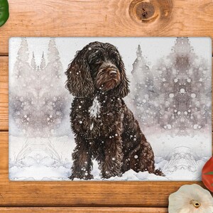 May include: A rectangular chopping board with a watercolour illustration of a brown dog in a snowy scene. The dog has long, curly fur and sits in the snow. The background depicts a snowy forest with falling snowflakes.