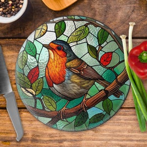 May include: Circular glass chopping board featuring a stained-glass style robin design. The bird has orange, red, and blue plumage, set against a backdrop of green leaves and light blue panes. A stainless steel knife and fresh vegetables are placed nearby.