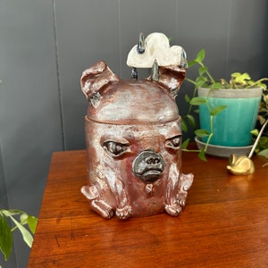 Może przedstawiać: Ceramiczna figurka chihuahua z chmurą na głowie. Figurka jest pomalowana w odcieniach brązu, czerni i bieli. Chihuahua ma ponury wyraz twarzy.