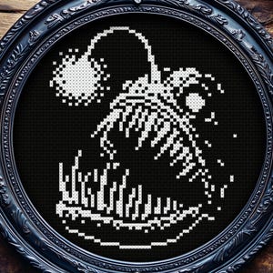 May include: A black framed cross-stitch artwork depicting a white anglerfish on a black backdrop. The anglerfish has a large mouth with sharp teeth and a glowing lure. The frame is ornate and black, set against a dark wood background.