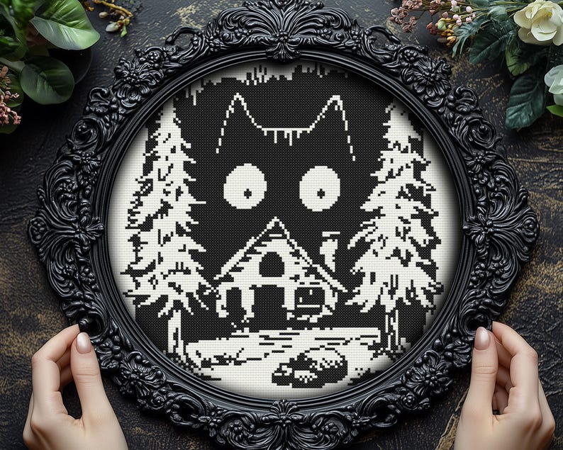 May include: Black and white cross-stitch embroidery of a cat's face with large eyes, a small house, and two pine trees. The embroidery is framed in a black ornate frame.