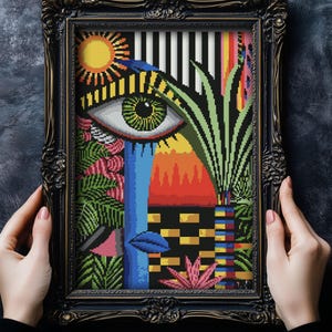 Puede incluir: Una obra de arte de punto de cruz enmarcada que presenta un diseño abstracto colorido con un ojo grande, formas geométricas y plantas tropicales. La obra está enmarcada en un marco de madera negro con detalles dorados.