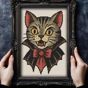 May include: Cross-stitch art of a cat with a vampire theme, framed in a black ornate frame. The cat has yellow eyes, a red bow tie, and a black cape. The cat's fur is gray and tan with black stripes.