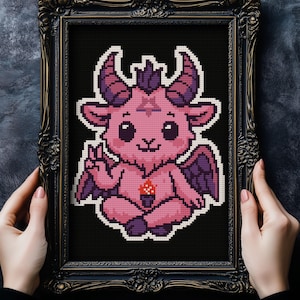May include: A framed cross-stitch featuring a pink, cartoonish Baphomet with purple horns, wings, and a pentagram. The artwork is set against a black background and framed in an ornate, dark-colored frame.