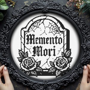 Könnte beinhalten: Schwarz gerahmte Kreuzstichkunst mit den Worten "Memento Mori" auf einem Grabstein-Design. Das Kunstwerk zeigt Rosen und Spinnweben. Der Rahmen ist verziert.