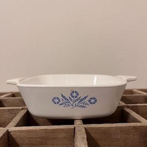 May include: White rectangular casserole dish with blue floral design. The dish has two handles and is sitting on a wooden surface. The floral design includes three flower clusters and leaves.