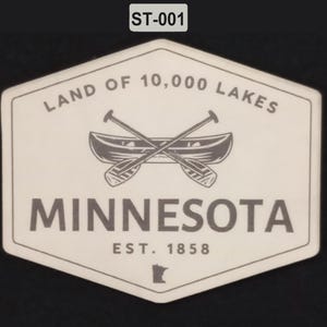 Op de afbeelding: Een zeshoekige sticker met een witte achtergrond en donkergrijze tekst en afbeeldingen. De tekst luidt LAND OF 10,000 LAKES, MINNESOTA en EST. 1858. In het midden staat een kano met twee gekruiste peddels, met daaronder de omtrek van de staat.