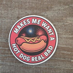Può includere: Un adesivo rotondo con bordo rosso e la scritta bianca "MAKES ME WANTA HOT DOG REAL BAD". Il centro presenta un'illustrazione di hot dog e fuochi d'artificio.