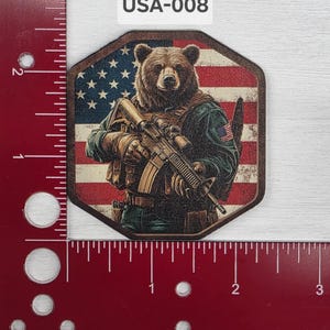 Puede incluir: Un oso marrón con uniforme militar y sosteniendo un rifle, de pie frente a una bandera estadounidense. La imagen está etiquetada como "USA-008".