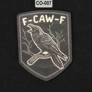 Può includere: Toppa a forma di scudo con un'illustrazione di un corvo. Il corvo è appollaiato su un ramo, con la scritta "F-CAW-F" sopra. La toppa ha una combinazione di colori grigio e bianco, con la scritta "CO-007" nell'angolo in alto a sinistra.