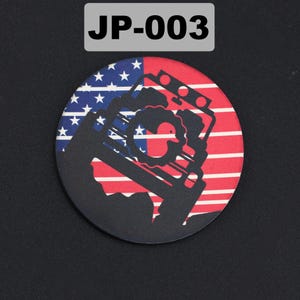 May include: A circular badge featuring a black silhouette of a Jeep-style vehicle with a rubber duck icon centered on the grille. The background displays the red, white, and blue American flag pattern. The text JP-003 is printed above the badge.