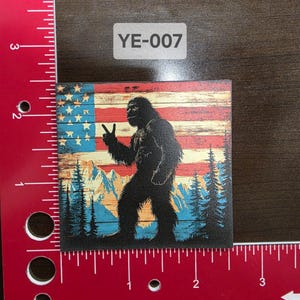 Può includere: Un adesivo quadrato con una silhouette di Bigfoot davanti a uno sfondo di bandiera americana. Il Bigfoot sta facendo un segno di pace con la mano sinistra. Il testo "YE-007" è stampato nell'angolo in alto a destra dell'adesivo.