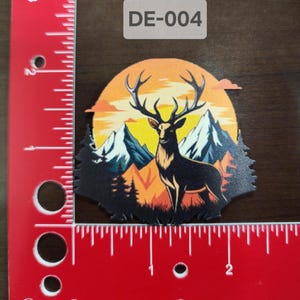 Puede incluir: Una pegatina troquelada con un ciervo majestuoso con grandes cuernos, sobre un fondo de montañas y un atardecer. La pegatina tiene un contorno negro y el texto "DE-004" en un rectángulo blanco.