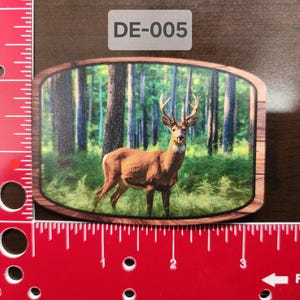 Puede incluir: Una placa de madera con marco marrón y una imagen impresa de un ciervo de cola blanca de pie en un bosque. El texto "DE-005" está impreso en la esquina superior derecha de la placa.