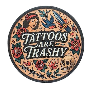 Peut inclure: Sous-verre rond, bordé de noir, avec un motif de style tatouage sur fond beige. Le motif comprend une femme, une hirondelle, un poignard, des roses, des flammes, un crâne et le texte "Tattoos are Trashy."